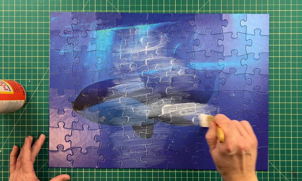 Applying puzzle glue with a brush across the surface of a completed blue jigsaw puzzle