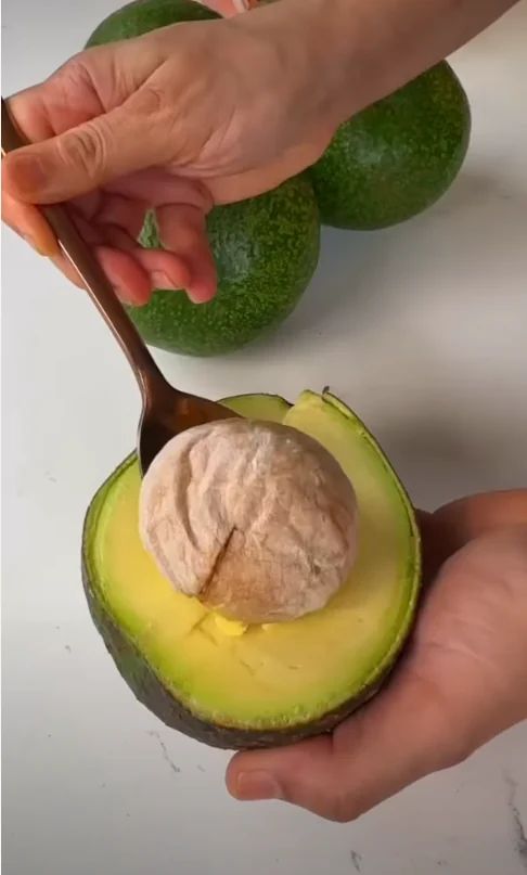 Ripe avocado cut in half with its large seed being lifted out using a spoon.
