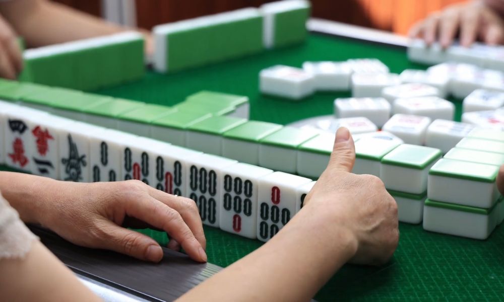 Close-up of players' hands and mahjong tiles arranged on a green gaming table.