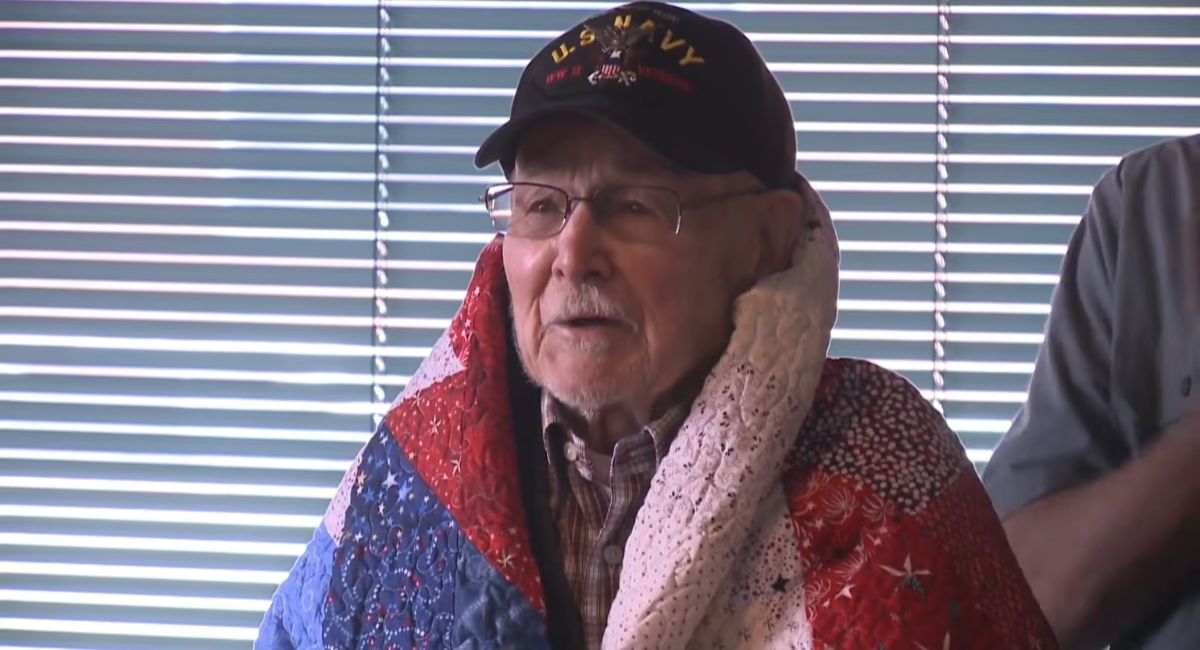 Elderly U.S. Navy veteran wrapped in a patriotic handmade quilt during a recognition ceremony.