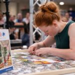 Young woman assembling a 500-piece jigsaw puzzle during a speed puzzling competition with spectators in the background.