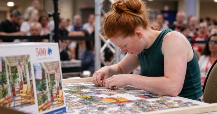 Young woman assembling a 500-piece jigsaw puzzle during a speed puzzling competition with spectators in the background.