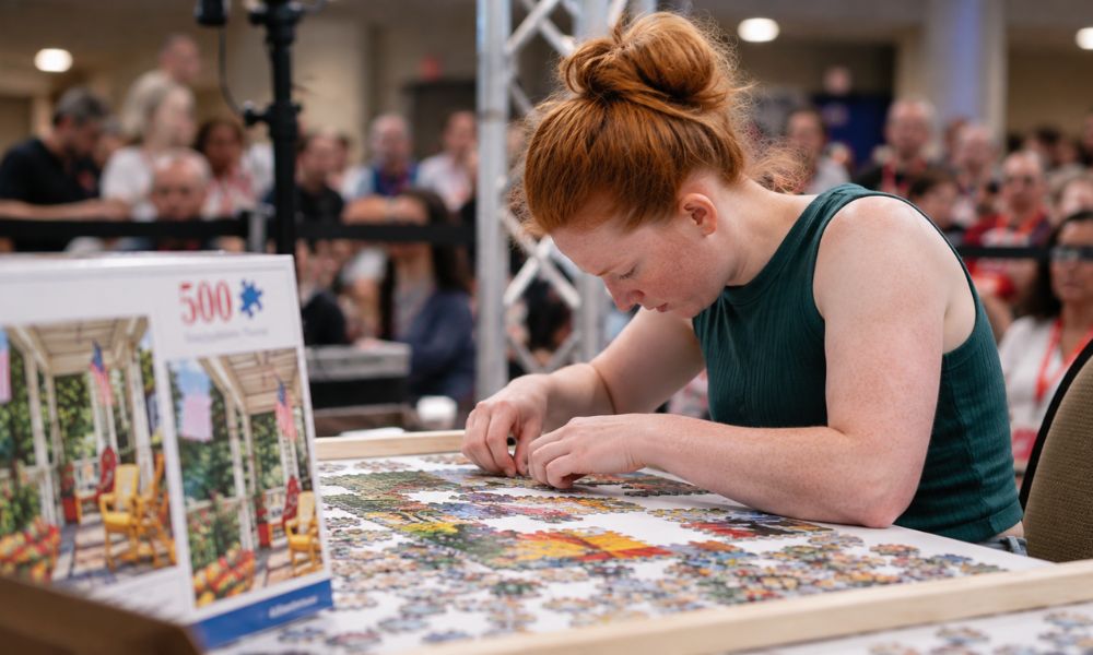 Young woman assembling a 500-piece jigsaw puzzle during a speed puzzling competition with spectators in the background.