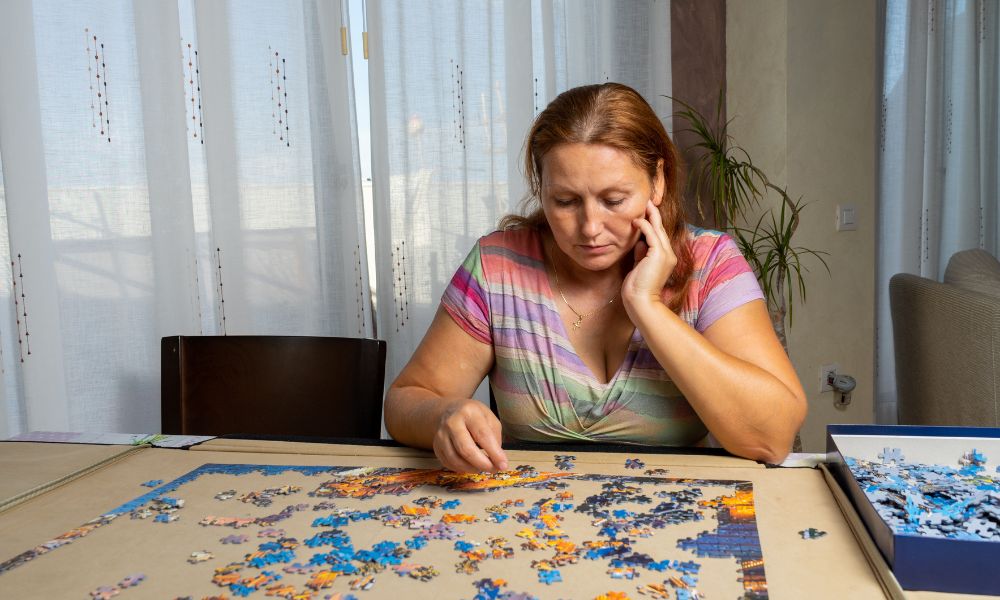 Woman assembling a colorful jigsaw puzzle at home with loose pieces spread across a table.