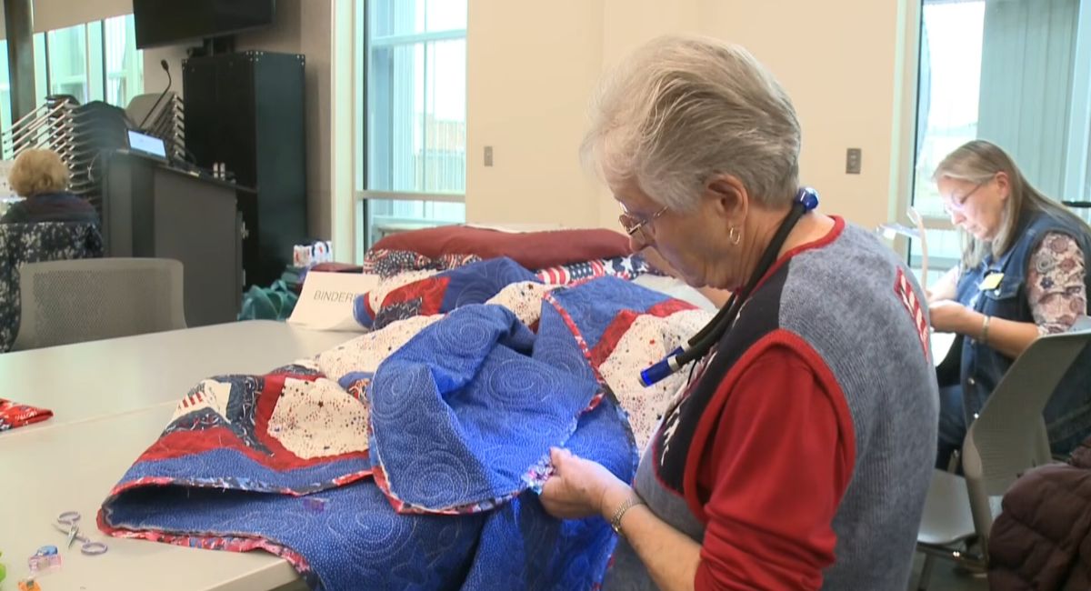 Elderly woman working on a red, white, and blue handmade quilt at a community quilting workshop.