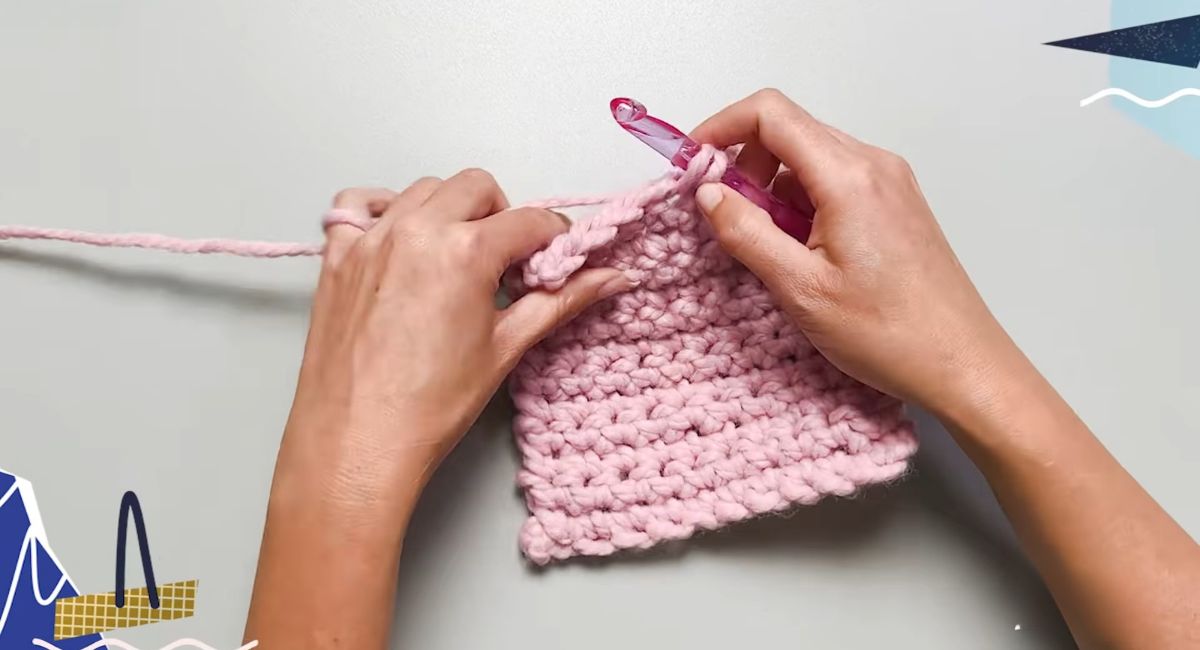 Hands crocheting pink yarn with a crochet hook, showing the formation of stitches.