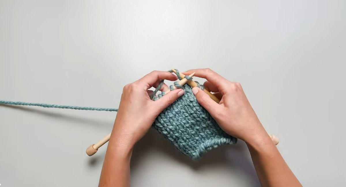 Hands knitting a blue-green fabric with wooden knitting needles.